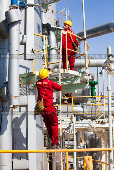 Two Asian maintenance workers in yellow hardhats and red work wear on distillation column of oil refinery plant on blue sky background. CNPC-AMG. company. (2012 год). Редакционное фото, фотограф Alexey Rezvykh / Фотобанк Лори