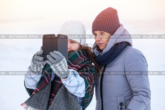 Two happy women friends sharing a smart phone after photo self shooting in cold winter lake shore. Стоковое фото, фотограф Кекяляйнен Андрей / Фотобанк Лори
