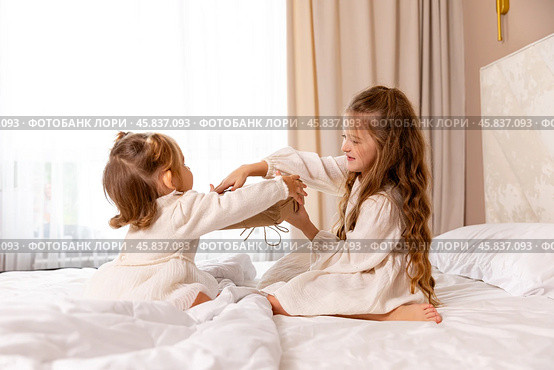 Two sisters wearing white dresses, sitting on a bed with white bedding, pulling big wrapped gift box, fighting, cannot share a present on the holiday morning at home. Стоковое фото, фотограф Ирина Михайличенко / Фотобанк Лори