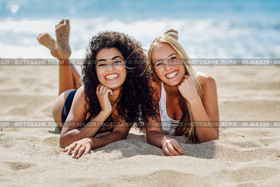 Two young women with beautiful bodies in swimwear on a tropical beach. Funny caucasian and arabic females wearing black and white swimsuits lying on the sand on the beach. Two young women with beautiful bodies in swimsuit on a tropical beach. Стоковое фото, агентство Ingram Publishing / Фотобанк Лори