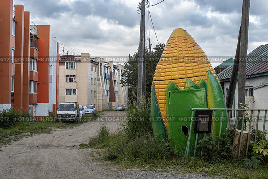 Улица провинциального города. Старый ларек в виде большого початка кукурузы. Автомобили припаркованы возле зданий на обочине грунтовой дороги. Петропавловск-Камчатский, Камчатский край, Россия. Редакционное фото, фотограф Андрей Степанов / Фотобанк Лори