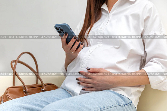 Unrecognizable pregnant woman using a smartphone and gently touching her belly while relaxing on couch in hospital. Happy young expectant mother. Family planning and IVF centers concept. High quality. Стоковое фото, фотограф Ирина Михайличенко / Фотобанк Лори