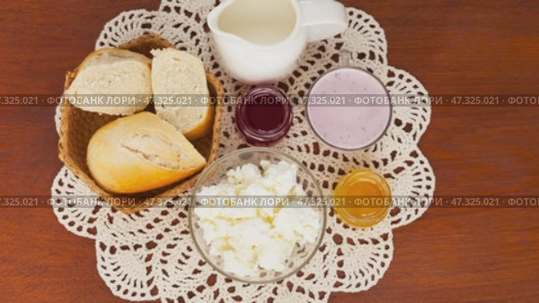 Video of delicious country breakfast with white bread and fresh dairy products - cottage cheese, yogurt, milk on hand-knitted lace napkin on wooden table. Rustic still life. Flat lay, close-up, top view. Стоковое видео, видеограф Виктория Катьянова / Фотобанк Лори