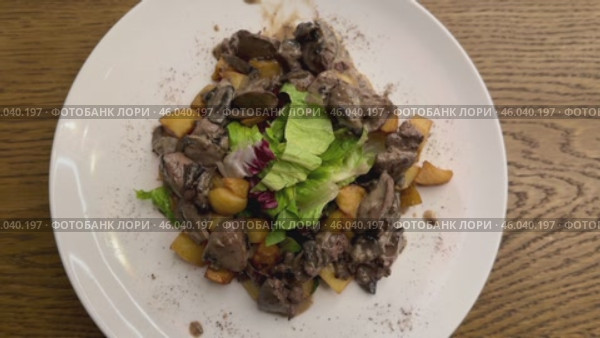Warm salad with chicken liver in cream sauce with fried potatoes, mushrooms and mixed green salad in a white porcelain plate on a brown wooden table in an elite restaurant. Стоковое видео, видеограф worker / Фотобанк Лори