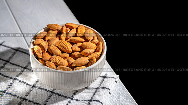 White bowl brimming with almonds resting on a checkered cloth atop a wooden table, creating a cozy, inviting atmosphere against a dark background. Perfect for highlighting healthy snacking. Стоковое фото, фотограф Ольга Ильина / Фотобанк Лори