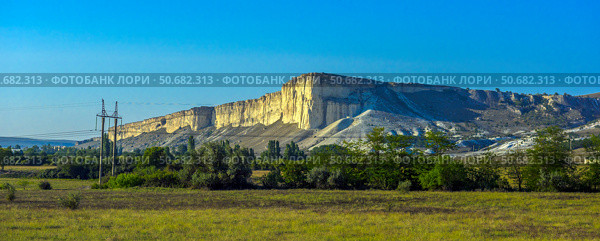 White Rock (Ak-Kaya) in Crimea is a monumental limestone formation with vertical cliffs, towering over a green valley. The rocky massif is flooded with sunlight, emphasizing the texture and light cream shade of the limestone. In the foreground are meadows and trees, creating a picturesque natural contrast with the majestic relief of the mountains. The sky is clear, bright blue, which adds a sense of space and grandeur to the frame. Стоковое фото, фотограф Анна Кривицкая / Фотобанк Лори