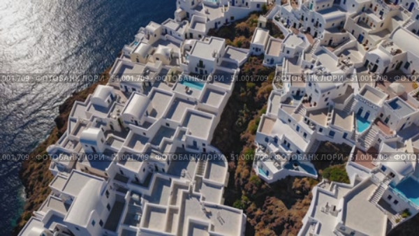 Whitewashed architecture cascading down volcanic cliffs of Santorini, revealing pristine hotels, swimming pools, and breathtaking views across sparkling Aegean waters. Стоковое AI-видео, аниматор Данил Руденко / Фотобанк Лори