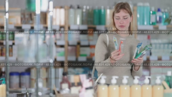 Woman in a jumper holds packs of toothbrushes with different levels of bristles for sensitive gums. Customer chooses dental hygiene accessories in a pharmacy. Стоковое видео, видеограф Яков Филимонов / Фотобанк Лори