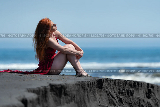 Woman in red long dress with raised hem and bare legs sitting on black sand on beach, hugging knees. Стоковое фото, фотограф А. А. Пирагис / Фотобанк Лори