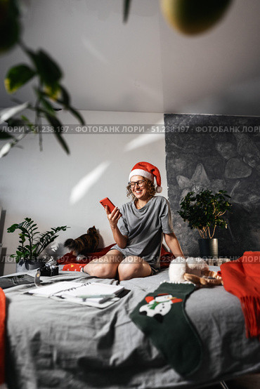 Woman in Santa hat enjoys Christmas at home, using smartphone for communication and social media. With help of modern technology, celebrates Christmas by sharing holiday moments and staying connected. Стоковое фото, фотограф Енваев Андрей / Фотобанк Лори