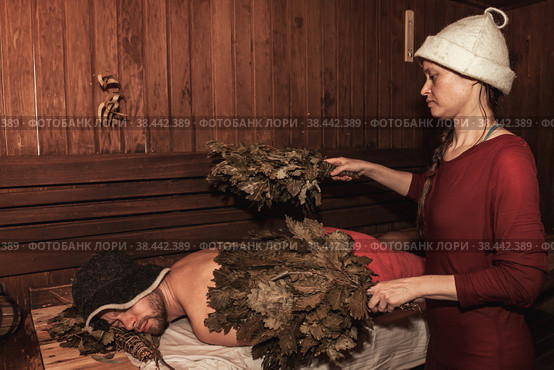 Woman in traditional dress prepare soak bath brooms in traditional steam sauna banya for man on bench in spa complex. Russian bathhouse. Wellness, self care, healthy concept. Copy text space. Стоковое фото, фотограф Aleksandr Golubev / Фотобанк Лори