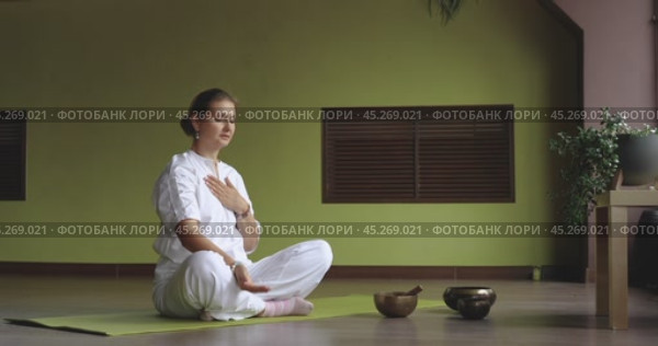 Woman practicing kundalini yoga, doing breathing practice raising arm up sharply throwing it behind head, sitting in white comfortable clothes in lotus position in room. Стоковое видео, видеограф Сергей Чайко / Фотобанк Лори