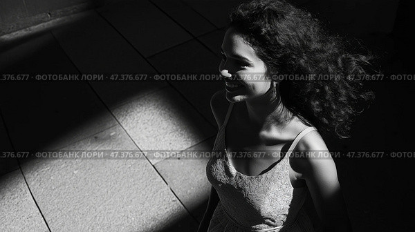 Woman walking in a sunlit corridor with shadows at noon. Женщина с вьющимися волосами улыбается, проходя по светлому коридору, наполненному контрастными тенями. Солнечный свет придает сцене глубину, пока она наслаждается моментом. Стоковое AI-изображение, AI-художник Katsiaryna Repich / Фотобанк Лори