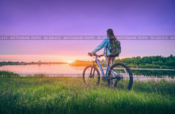 Woman with bicycle near the river at sunset in spring. Landscape with sporty girl with backpack riding a mountain bike, dirt road, green grass, water, purple sky in summer. Sport and travel. Cycle. Стоковое фото, агентство Ingram Publishing / Фотобанк Лори