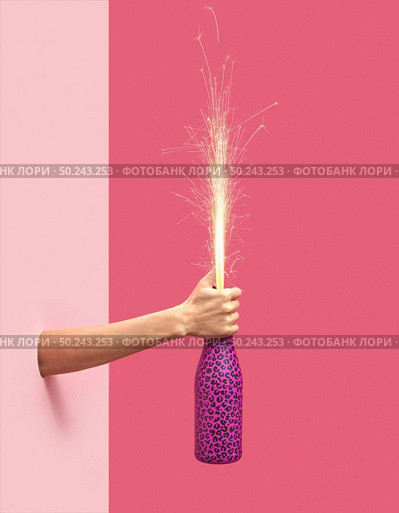 Woman’s hand holds painted champagne bottle with holiday firecracker on a duotone pink background, copy spase. Christmas concept.. Holiday firework in a painted bottle in a female’s hand. Стоковое фото, агентство Ingram Publishing / Фотобанк Лори