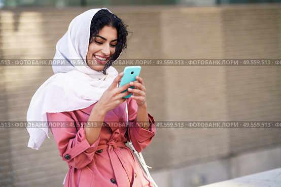 Young Arab woman wearing hijab headscarf texting message with her smartphone in urban background.. Young Arab woman wearing hijab texting message with her smartphone. Стоковое фото, агентство Ingram Publishing / Фотобанк Лори