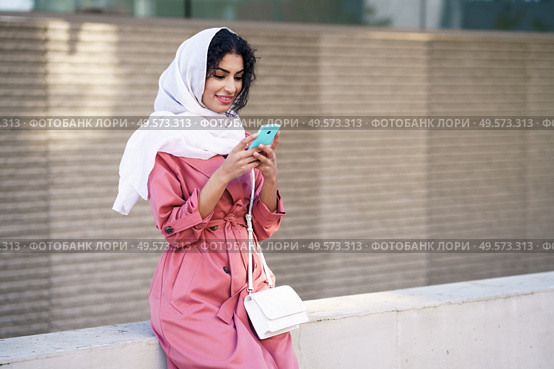 Young Arab woman wearing hijab headscarf texting message with her smartphone in urban background.. Young Muslim woman wearing hijab texting message with her smartphone. Стоковое фото, агентство Ingram Publishing / Фотобанк Лори