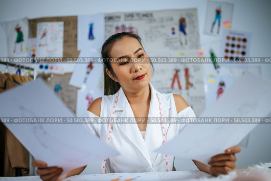 Young Asian fashion designer or tailor female checking on clothes sketch in hand, Blured dress and beautiful sketch on board in background. Стоковое фото, агентство Ingram Publishing / Фотобанк Лори