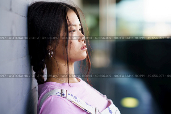 Young Asian female in casual clothes with braid leaning on wall and looking away on city street in daytime. Asian woman leaning on wall. Стоковое фото, агентство Ingram Publishing / Фотобанк Лори
