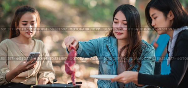 Young asian woman cooking and her friend enjoy to make the meal in pan, They are talk and laugh with fun together while camping in nature park. Стоковое фото, агентство Ingram Publishing / Фотобанк Лори