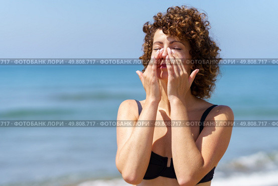 Young female in bikini applying sunscreen lotion on face with hands while standing near waving sea during summer weekend. Woman smearing sunblock cream on face near sea. Стоковое фото, агентство Ingram Publishing / Фотобанк Лори