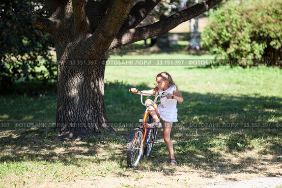 Young girl trying to ride her bicycle in summer park on meadow with green grass. Стоковое фото, фотограф Кекяляйнен Андрей / Фотобанк Лори