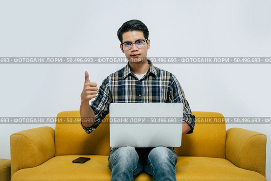 Young handsome man wearing eyeglasses with new normal, sitting on sofa with thump up, Heworks from home on laptop computer while self-isolation and quarantine covid-19 coronavirus outbreak. Стоковое фото, агентство Ingram Publishing / Фотобанк Лори