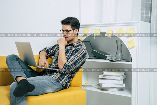 Young handsome man wearing eyeglasses with new normal, sitting on sofa and works from home on laptop computer while self-isolation and quarantine covid-19 coronavirus outbreak. Стоковое фото, агентство Ingram Publishing / Фотобанк Лори