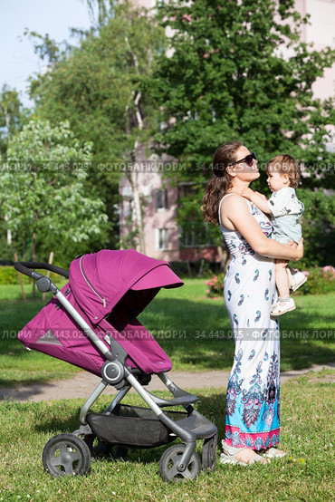 Young mother in summer light dress holding her toddler daughter in arms, woman standing close to baby stroller on green lawn. Стоковое фото, фотограф Кекяляйнен Андрей / Фотобанк Лори
