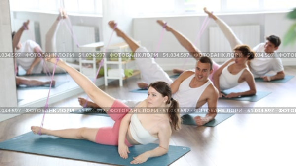 Young sporty women and men doing pilates exercises with fitness resistance bands lying on side stretching legs in light gym. Стоковое видео, видеограф Яков Филимонов / Фотобанк Лори