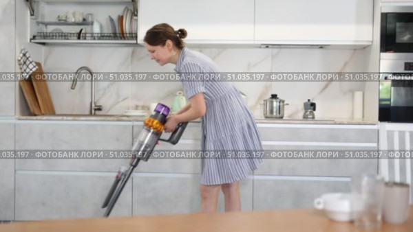 Young woman in homemade blue dress doing household chores. Experienced housewife with vacuum cleaner furbish cleanse carpeting synthetic covering near dining table in kitchen. Стоковое видео, видеограф Яков Филимонов / Фотобанк Лори