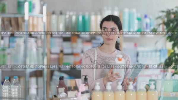 Young woman in sweater standing near racks of medicines and pills and choosing moisturizer with collagen and body lotion at pharmacy. Стоковое видео, видеограф Яков Филимонов / Фотобанк Лори