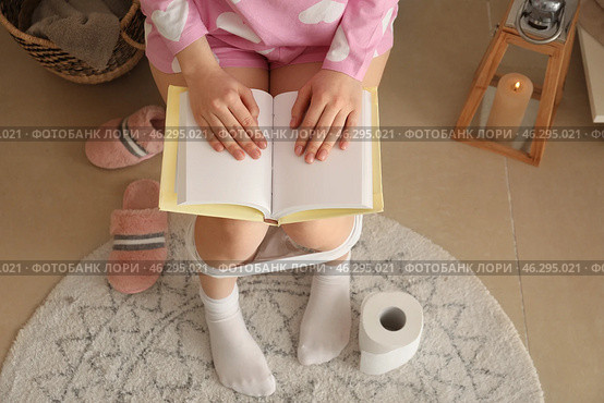Young woman reading book while sitting on toilet bowl in restroom. Стоковое фото, фотограф Pixel_shot / Фотобанк Лори