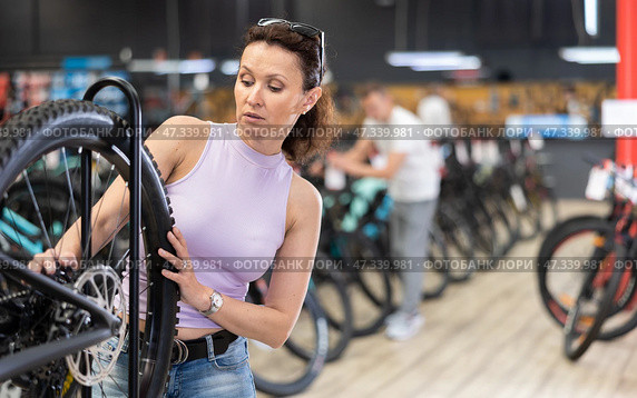Young woman selects bicycle in sporting goods store, checking wheel quality. Стоковое фото, фотограф Яков Филимонов / Фотобанк Лори