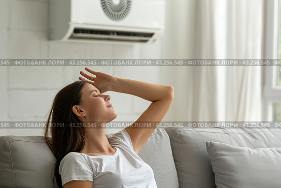 Young woman suffering during a heat heat sitting under air conditioner at home resting on a sofa. Exhausted girl feeling sweaty switching on modern AC system. Hot summer. Fresh clean air in apartment. Стоковое AI-изображение, AI-художник Марина Демидюк / Фотобанк Лори