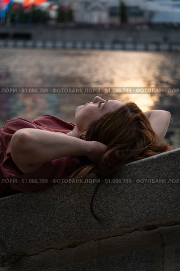 Young woman with red hair in a burgundy dress relaxing by the riverbank at sunset, with warm golden reflections shimmering on the water. Стоковое фото, фотограф Софья Соломонова / Фотобанк Лори