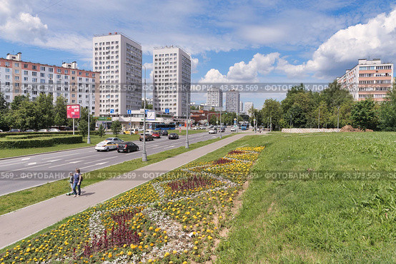 Женщина с ребенком солнечным летним днем идут по тротуару вдоль Центрального проспекта подмосковного города Зеленограда. Зеленый газон с цветами, жилые дома, голубое небо, белые облака. (2024 год). Стоковое фото, фотограф Сергей Никифоров-Гордеев / Фотобанк Лори